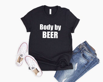 Op de afbeelding: Een zwart T-shirt met witte tekst die "Body by BEER" zegt. Het shirt ligt plat op een wit oppervlak met een paar witte sneakers en een blauwe jeans.