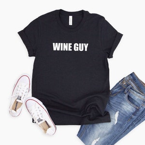 May include: A black t-shirt with the text "WINE GUY" printed in white on the front. The shirt is paired with blue jeans and white sneakers.