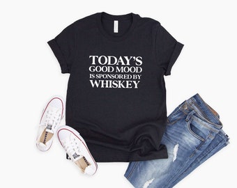 May include: A black t-shirt with white text that reads "TODAY'S GOOD MOOD IS SPONSORED BY WHISKEY". The shirt is paired with blue jeans and white sneakers.
