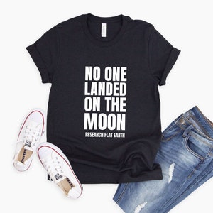 May include: A black t-shirt with white text that reads "NO ONE LANDED ON THE MOON RESEARCH FLAT EARTH". The shirt is laid flat on a white background with a pair of white sneakers and blue jeans.