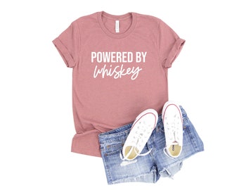 Peut inclure: Un t-shirt rose avec le texte "POWERED BY whiskey" en lettres blanches. Le t-shirt est associé à un short en jean et des baskets blanches.