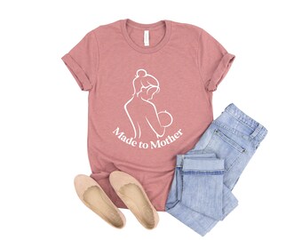 May include: A dusty rose t-shirt with a white outline of a woman holding a baby and the text "Made to Mother".