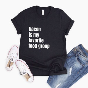 Op de afbeelding: Zwart T-shirt met witte tekst die "bacon is my favorite food group" zegt. Het shirt wordt gecombineerd met een blauwe jeans en witte sneakers.