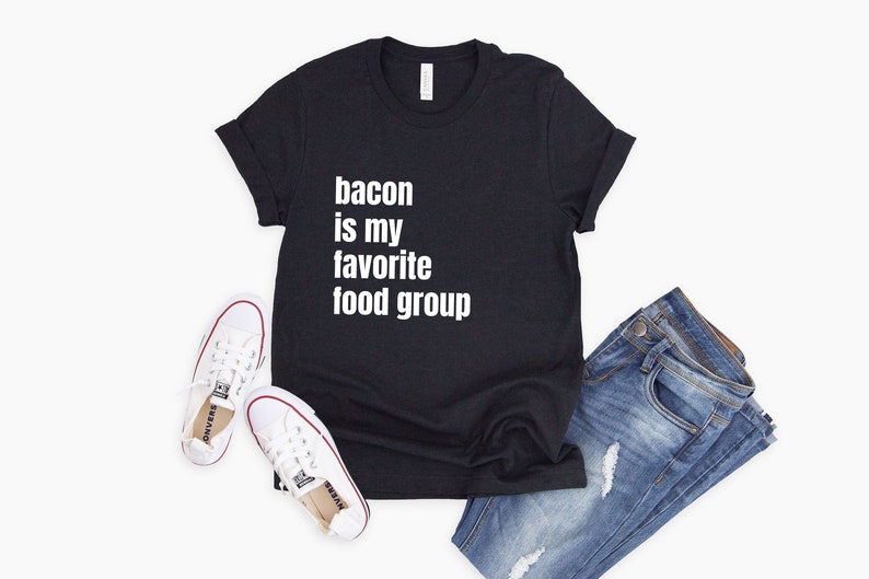 Op de afbeelding: Zwart T-shirt met witte tekst die "bacon is my favorite food group" zegt. Het shirt wordt gecombineerd met een blauwe jeans en witte sneakers.