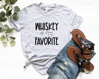 Peut inclure: Un t-shirt gris avec le texte "WHISKEY is my FAVORITE" imprimé en noir. Le t-shirt est posé sur une surface en bois blanc avec une paire de sandales marrons et un jean bleu.