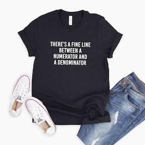 May include: A black t-shirt with white text that reads "There's a fine line between a numerator and a denominator". The shirt is laying on a white surface with a pair of white sneakers and blue jeans.