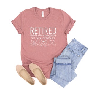 以下が含まれることがあります： 「RETIRED UNDER NEW MANAGEMENT SEE CATS FOR DETAILS」の白文字が入ったダスティローズのTシャツ。ライトブルーのジーンズとベージュのフラットシューズとコーディネートされています。Tシャツには猫のグラフィックが描かれています。