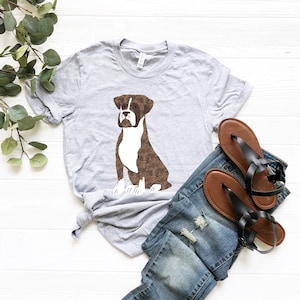 May include: A gray t-shirt with a brown and white boxer dog illustration. The dog is sitting and looking to the right. The t-shirt is on a white surface with a pair of brown sandals and blue jeans.
