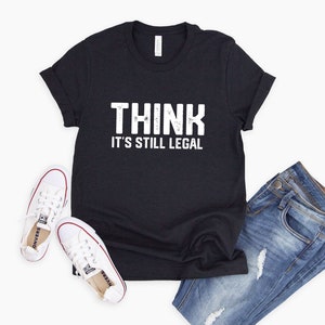 May include: Black t-shirt with the text "THINK IT'S STILL LEGAL" in white. The shirt is paired with light blue denim jeans and white canvas trainers with red accents.