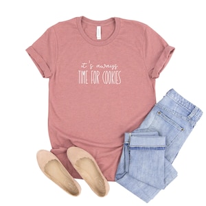 May include: A pink t-shirt with the text "it's always time for cookies" in white lettering. The shirt is paired with blue jeans and beige flats.