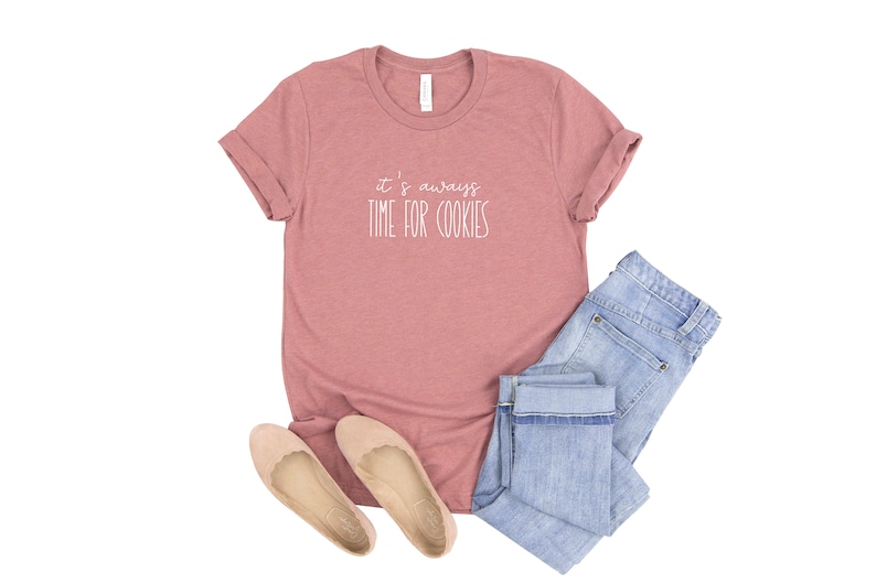 May include: A pink t-shirt with the text "it's always time for cookies" in white lettering. The shirt is paired with blue jeans and beige flats.
