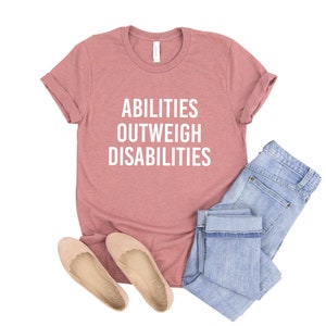 May include: A dusty rose t-shirt with the text "ABILITIES OUTWEIGH DISABILITIES" in white lettering. The shirt is paired with blue jeans and beige flats.