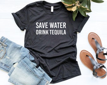 Puede incluir: Camiseta negra con texto blanco que dice "Save Water Drink Tequila". La camiseta está puesta sobre una superficie blanca con un par de sandalias marrones y un par de pantalones vaqueros azules.