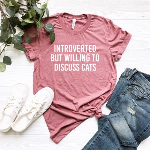 May include: A maroon t-shirt with the text "Introverted but willing to discuss cats" in white lettering.