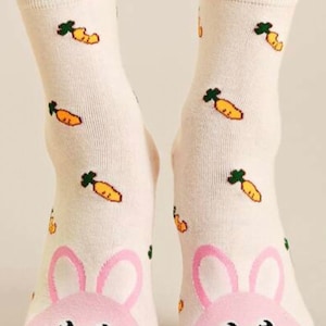 Puede incluir: Un par de calcetines blancos hasta el tobillo con una cara de conejo rosa en los dedos y un patr&oacute;n de zanahorias verdes sobre un fondo blanco.