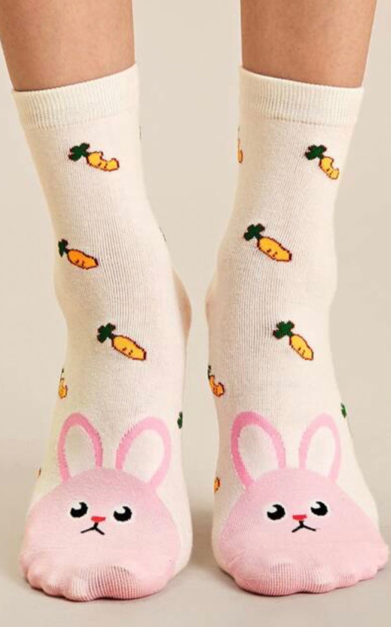 Puede incluir: Un par de calcetines blancos hasta el tobillo con una cara de conejo rosa en los dedos y un patr&oacute;n de zanahorias verdes sobre un fondo blanco.