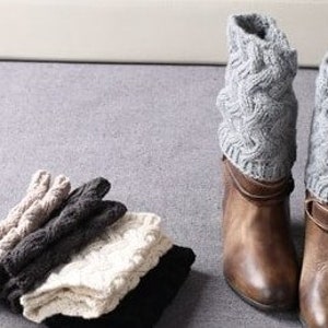 May include: Four pairs of knitted boot cuffs in beige, black, gray, and white. The cuffs are folded and stacked on top of each other. Two pairs of brown leather boots with gray knitted boot cuffs are shown in the background.