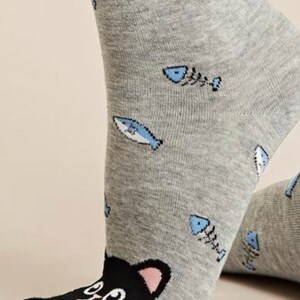 Puede incluir: Calcetines grises de tobillo con una cara de gato negro en la punta y un patr&oacute;n de peces azules y huesos.