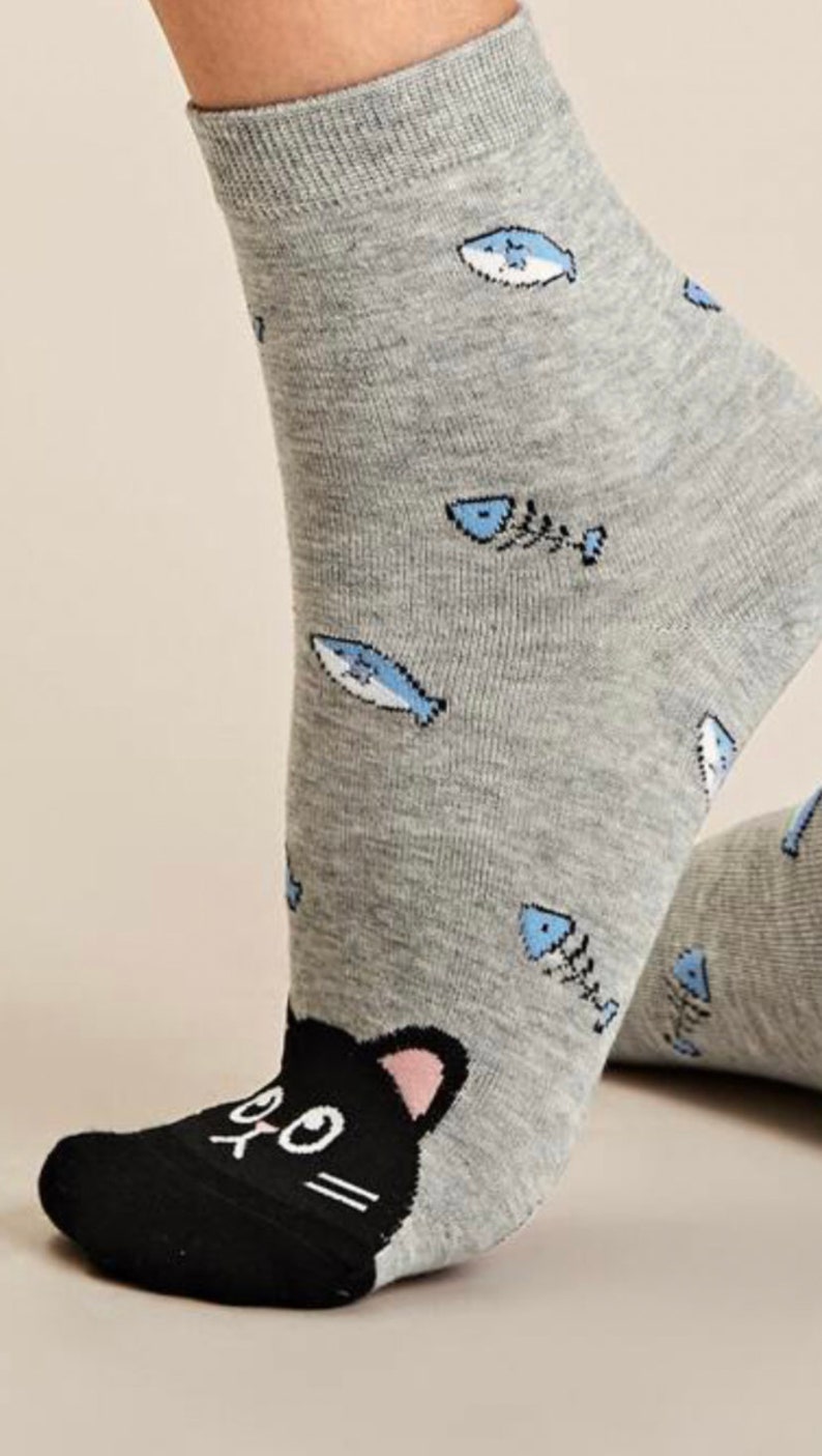 Puede incluir: Calcetines grises de tobillo con una cara de gato negro en la punta y un patr&oacute;n de peces azules y huesos.