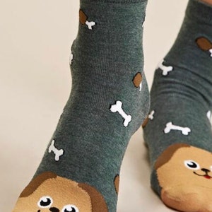 Puede incluir: Un par de calcetines de tobillo con fondo verde y un dise&ntilde;o de cara de perro en los dedos. Los calcetines tambi&eacute;n presentan dise&ntilde;os en forma de hueso blanco y marr&oacute;n.