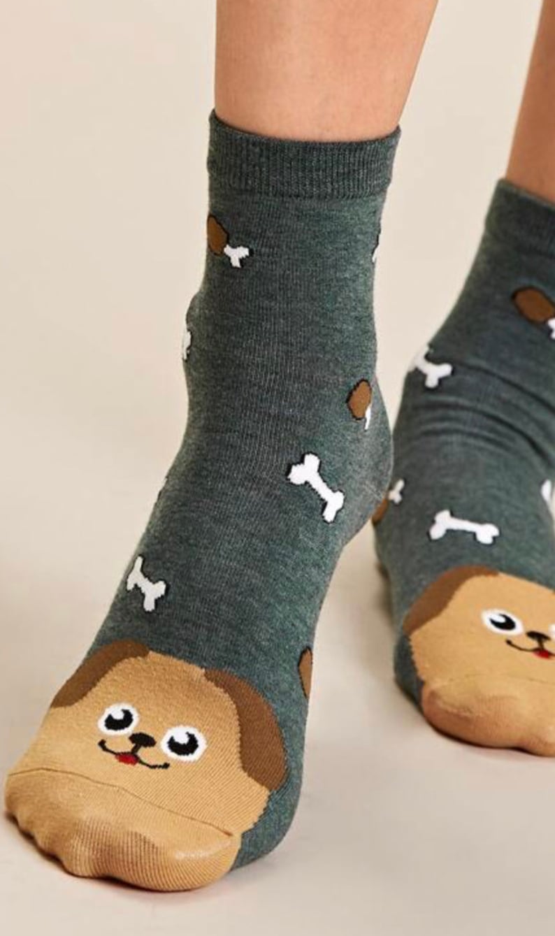 Puede incluir: Un par de calcetines de tobillo con fondo verde y un dise&ntilde;o de cara de perro en los dedos. Los calcetines tambi&eacute;n presentan dise&ntilde;os en forma de hueso blanco y marr&oacute;n.