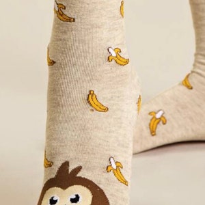 Puede incluir: Un par de calcetines de tobillo beige con una cara de mono marr&oacute;n en la punta y un patr&oacute;n de pl&aacute;tano amarillo.