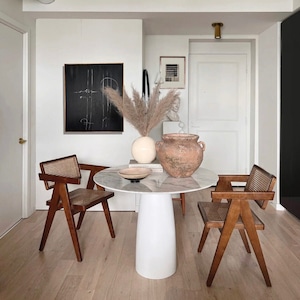 Pode incluir: Uma mesa redonda branca com uma base de pedestal branca, duas cadeiras de madeira marrom com assentos de vime trançado, um vaso de terracota grande com uma planta e uma imagem emoldurada preta na parede.