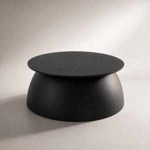 Pode incluir: Uma mesa de centro preta moderna com um tampo redondo e uma base arredondada. O tampo tem vários orifícios pequenos e espaçados uniformemente. A mesa é feita de um material preto fosco e está sobre um fundo neutro.