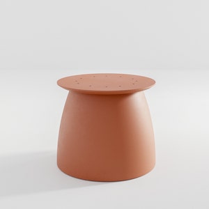 Pu&ograve; includere: Un tavolo rotondo a piedistallo di colore terracotta con una superficie liscia leggermente curva. Il tavolo presenta una serie di piccoli fori attorno al bordo superiore.