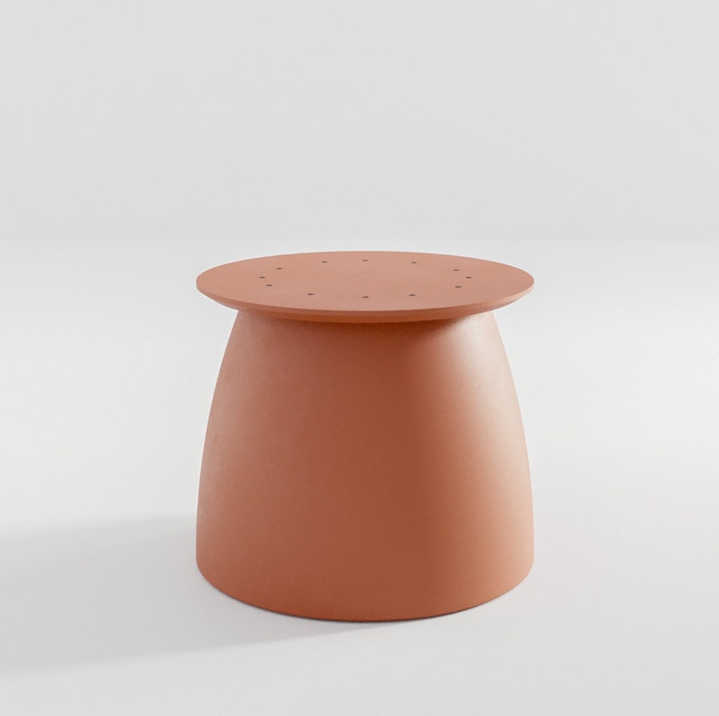 Pu&ograve; includere: Un tavolo rotondo a piedistallo di colore terracotta con una superficie liscia leggermente curva. Il tavolo presenta una serie di piccoli fori attorno al bordo superiore.