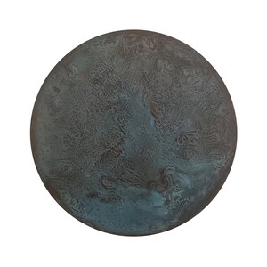 Pode incluir: Um objeto circular com uma superfície escura e desgastada, apresentando uma textura manchada. O acabamento exibe uma mistura de tons castanho-escuros, carvão e azul-petróleo oxidado, criando um padrão orgânico irregular na face redonda.