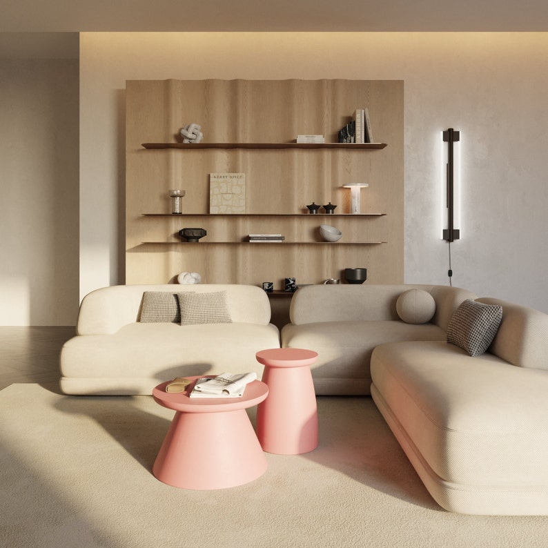 May include: A white sectional sofa with two pink side tables in a living room with a wooden shelf unit behind the sofa. The shelf unit has books, decorative objects, and a lamp on it.