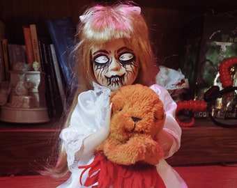 May include: A porcelain doll with blonde hair, wearing a white dress with lace trim, holds a brown teddy bear. The doll has dark eye makeup and a painted smile.