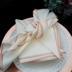 May include: An ivory napkin with rose gold trim, knotted on a white plate with a beaded edge, atop a rose gold charger plate. A stylish table setting for a special occasion.