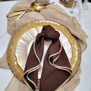 May include: A brown napkin with a gold trim is folded into a decorative knot and placed on a gold and white plate. The plate is on a beige tablecloth.