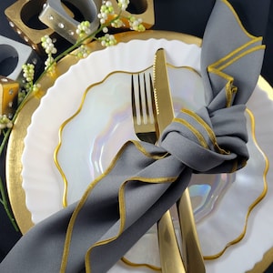 May include: A gray linen napkin with gold trim is tied around a gold fork and knife on a white and gold rimmed plate. The plate is on a gold charger.
