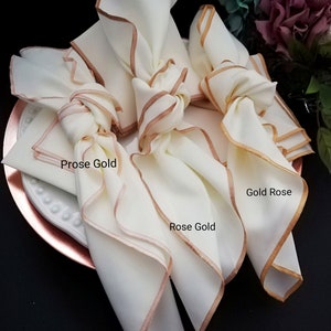 May include: Cream-coloured cloth napkins with rose gold trim, knotted and arranged on a rose gold charger plate. The napkins have the text "Prose Gold" and "Gold Rose" and "Rose Gold" printed on them. The napkins are part of a table setting.