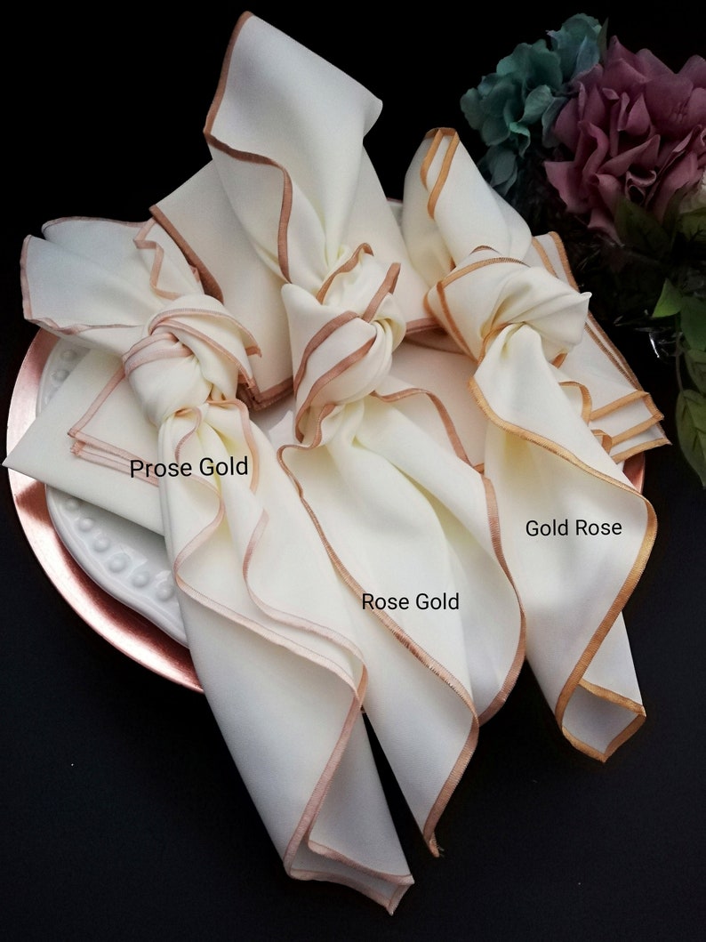 May include: Cream-coloured cloth napkins with rose gold trim, knotted and arranged on a rose gold charger plate. The napkins have the text "Prose Gold" and "Gold Rose" and "Rose Gold" printed on them. The napkins are part of a table setting.