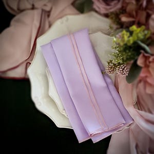 Pu&ograve; includere: Tovaglioli lavanda piegati con un bordo rosa chiaro, presentati su un piatto decorativo bianco. I tovaglioli sono lisci e sembrano essere fatti di un tessuto morbido. Composizioni floreali e tessuto drappeggiato sullo sfondo.