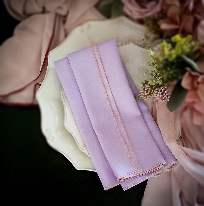 Pu&ograve; includere: Tovaglioli lavanda piegati con un bordo rosa chiaro, presentati su un piatto decorativo bianco. I tovaglioli sono lisci e sembrano essere fatti di un tessuto morbido. Composizioni floreali e tessuto drappeggiato sullo sfondo.