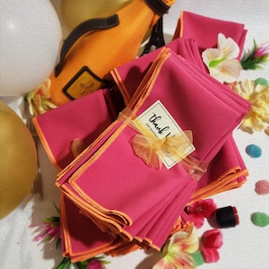 May include: A stack of pink napkins with orange trim, tied with a yellow ribbon and a tag that says "Thank You" in gold lettering. The napkins are surrounded by yellow and pink flowers and other decorative items.