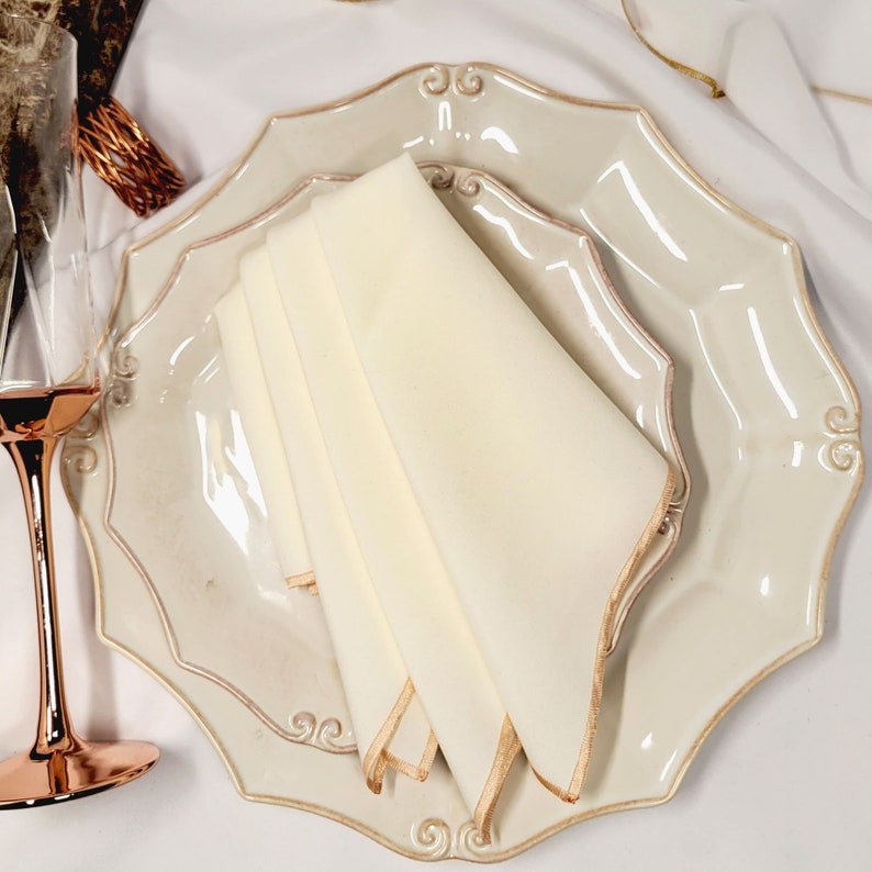 May include: A folded cream napkin sits on a decorative off-white plate with a scalloped edge and gold trim. The plate is part of a formal table setting. A champagne glass is visible in the background.