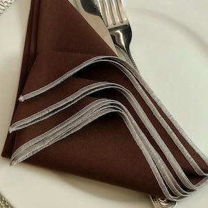 May include: A folded brown napkin with silver trim rests on a white plate, next to a knife and fork. The napkin is folded into a decorative shape, with the silver trim adding a touch of elegance to the table setting.