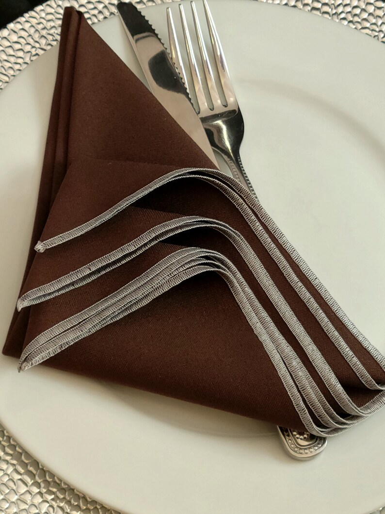 May include: A folded brown napkin with silver trim rests on a white plate, next to a knife and fork. The napkin is folded into a decorative shape, with the silver trim adding a touch of elegance to the table setting.