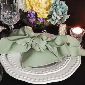 May include: A formal table setting featuring a light green napkin tied in a knot, on white plates with a beaded edge. Includes crystal glassware, pearl napkin rings, and a lit candle.