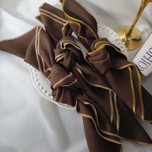 May include: Four brown cloth napkins with gold trim are knotted and arranged on a white plate. The napkins have a folded design with a contrasting gold border. A gold-stemmed wine glass is visible in the background.