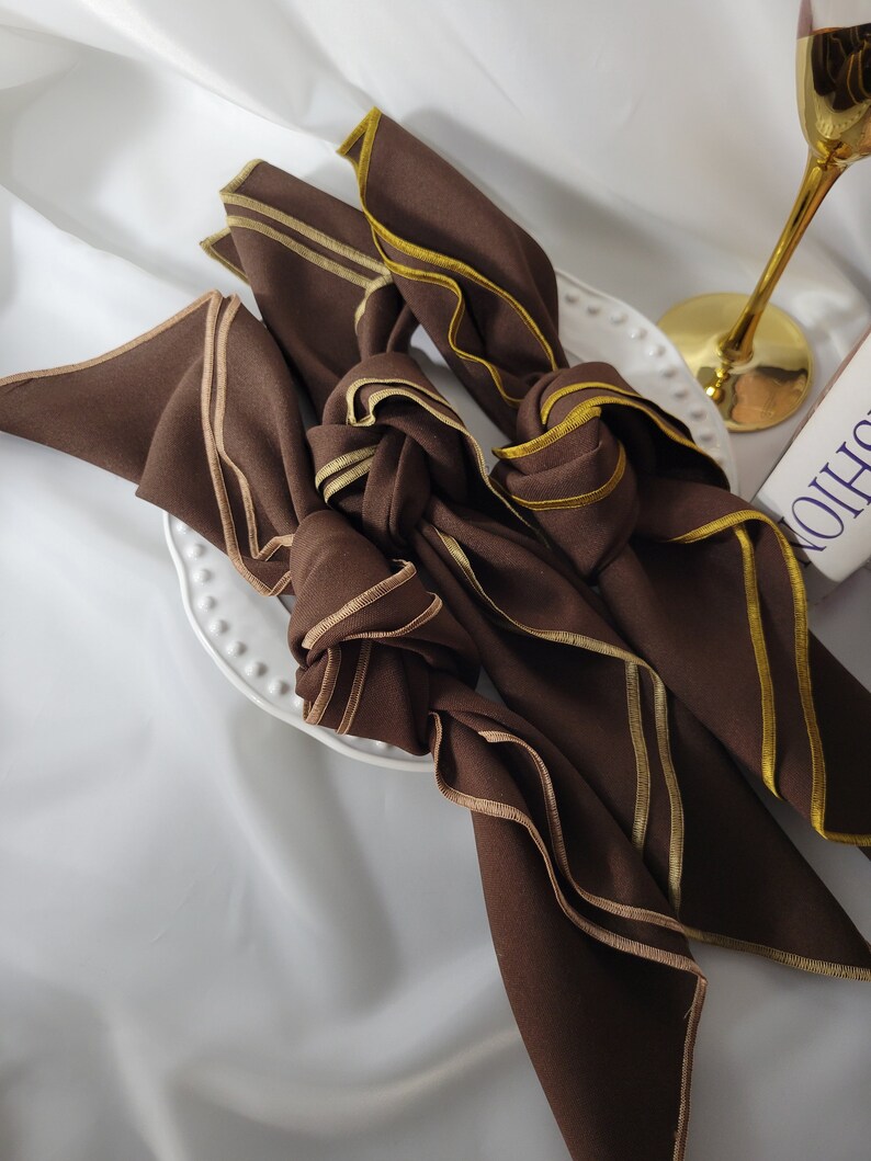 May include: Four brown cloth napkins with gold trim are knotted and arranged on a white plate. The napkins have a folded design with a contrasting gold border. A gold-stemmed wine glass is visible in the background.