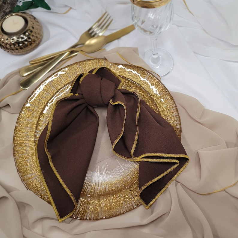 May include: A brown napkin with a gold trim is folded into a bow and placed on a gold and clear glass charger plate.