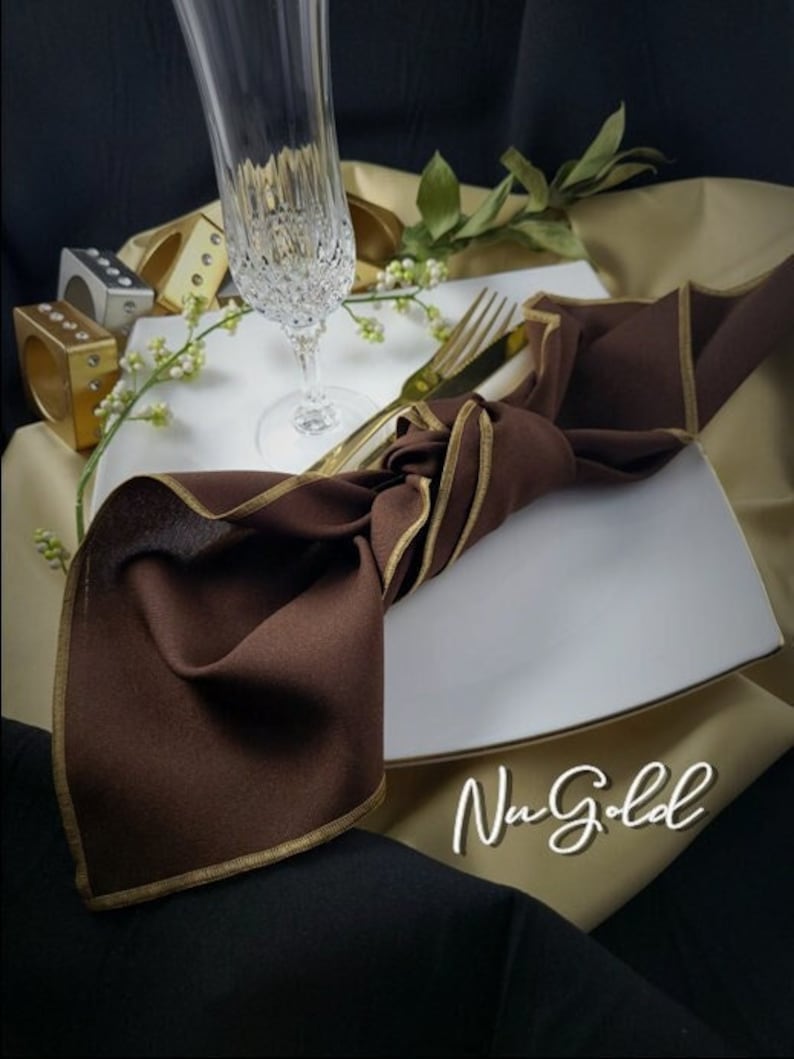 May include: A brown napkin with gold trim is knotted on a white square plate. A champagne flute, gold-colored silverware, and decorative ring holders are arranged on a beige and black background. The text "Nu Gold" is visible.