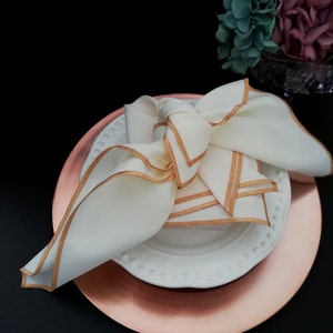 May include: A folded white linen napkin with gold trim sits on a white plate with a beaded edge, placed on a copper-coloured charger plate. The napkin is tied in a decorative knot. The setting is on a black surface.